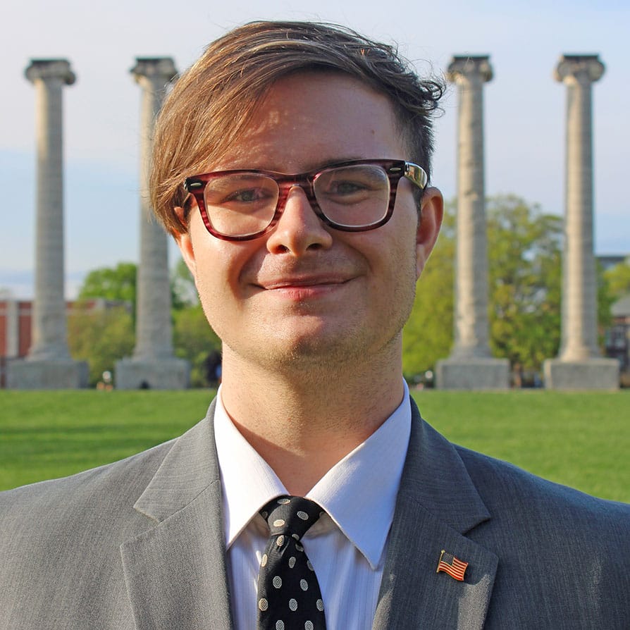 A headshot photograph of me. I am wearing a gray suit, a black tie, and glasses. The background is the six columns on MU's Francis Quadrangle.
